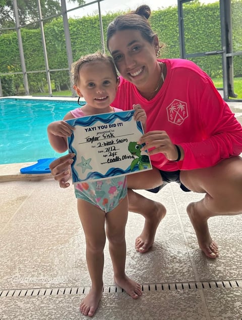 Woman and young child by a swimming pool holding a certificate together, both smiling at the camera