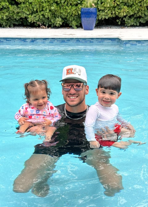 Adult man with two young children in a swimming pool, all smiling at the camera with lush green foliage visible in background