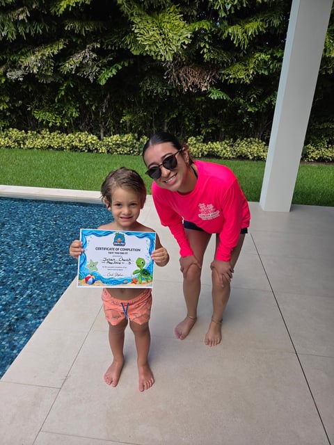 Adult and young child standing by a pool holding a swimming certificate, both smiling at the camera