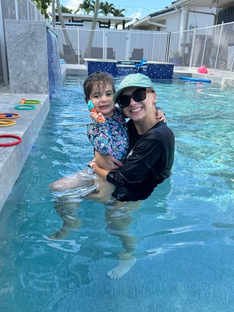 Adult holding young child in pool, both smiling at camera. Clear blue water, pool toys visible, residential setting with fence in background.