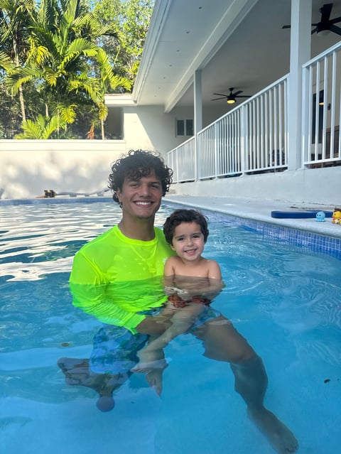 Two children smiling in a bright blue swimming pool on a sunny day with a white house and palm trees in the background